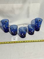 Five cobalt blue drinking glasses with white crescent moons and gold stars, arranged in a row on a white surface with measuring tape in front for size reference.