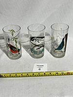 Three clear glass tumblers with colorful bird designs from Charley Harper, shown side by side with measuring tape for scale.