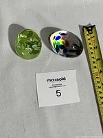Two art glass pieces placed on a white cloth next to a measuring tape showing size, one green with white floral inclusions, the other clear with multicolored spots.