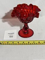 Front view of the ruby red compote showing its ruffled edge and pedestal base, next to a tape measure and lot card.