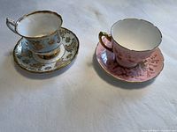 Two teacup and saucer sets on white cloth background; left is blue and gold, right is pink and gold