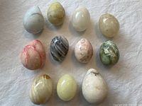 Eleven marble eggs of various colors and sizes displayed on white fabric, showing natural veining and patterns on each egg.