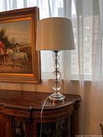 Full view of the crystal ball table lamp with white shade on wooden furniture near window and painting