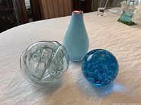 Clear glass knot, blue paperweight with bubbles, and blue glass vase with red rim arranged on a white cloth surface.