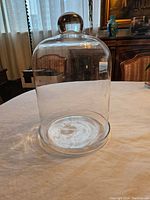 Large clear glass cloche with rounded dome top and knob handle placed on a pale tablecloth with a dark wood furniture background.
