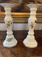 Pair of white ceramic candlesticks with floral hand-painted decoration, standing upright on a wooden surface in front of a framed painting.