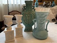 Photo of assorted blue and white ceramic vases and decorative pieces arranged on a table by window. Includes two small white urn-shaped pieces and two blue vases with handles and embossed floral details.