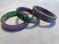 Three Chinese jade bracelets arranged horizontally showing carved floral and ornamental designs in green and purple jade.