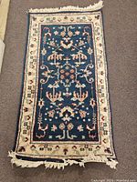 Full view of the small rectangular carpet showing the blue center with beige floral border and fringe wear.