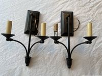 Photo showing a pair of traditional black metal wall sconces each with two arms. One sconce has a missing candle sleeve.
