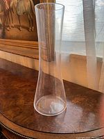 Tall narrow clear glass vase with slight flare at base, on wooden surface by window with curtain and painting background.