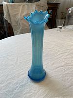 Photo showing a tall blue opalescent glass vase with a ruffled, flared rim and thick base, placed on a tablecloth with a plain background.