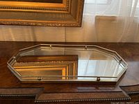 Octagonal mirrored tray on wooden surface reflecting gold frame painting and sheer curtains.