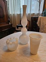Photo of three white vases displayed on a table in a room with natural light through curtains.