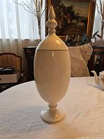White tall wooden decorative urn with lid, smooth white finish, pedestal base and pointed finial top.