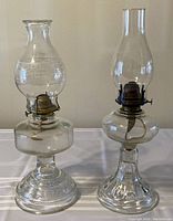 Two glass kerosene lamps on cloth surface, showing oil reservoirs, chimneys, and burner portions. Lamps are clear glass with metal burners and no oil inside.