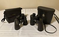 Photo of two pairs of binoculars on a white cloth surface. The Bushnell binoculars with a black soft case on the left and the Carl Schulz binoculars with a black hard case on the right.
