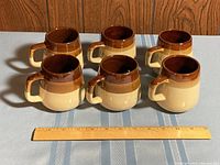 Photo showing six matching coffee mugs with a two-tone brown and beige color scheme arranged in two rows on a table with a wooden ruler in front for scale.