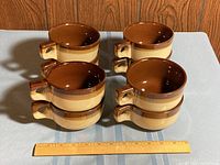 Six brown and beige onion soup bowls arranged in two stacks of three on a surface with a wooden ruler for scale.