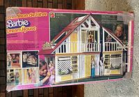 Front image of the original box showing pink and white framed Barbie Dream House with dolls in and around it, details of front facade, yellow door, and windows.