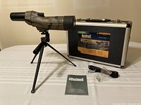 Photo showing Bushnell camo spotting scope on tripod, hard case, manual, cleaning cloth and strap on tabletop.