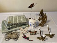 Butterfly-themed collection on white surface showing sage green tablecloth and napkins, multiple butterfly decor items including metal trivet and brass pieces, ceramic candy dish, hanging ornament, tealight holder and small Mothers Day decor