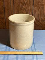 Cream-colored stoneware crock placed on a table with a wooden ruler in front for size reference. Crock has a smooth finish and no visible maker's mark or lid.