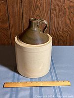 Photo showing two-tone vintage crock with handle, beige base and dark brown glazed top, placed behind a wooden ruler for size reference.