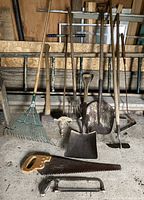 Photo showing an assortment of garden tools including rakes, shovels, hoes, an ax, and saws leaning against a wooden wall in a rustic environment.