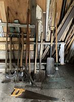Overview of garden tools including rakes, shovels, hoes, ax, saw arranged against a wooden backdrop