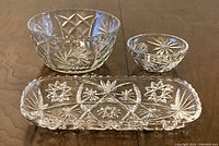 Three clear glass serving pieces on brown surface, including a large bowl, small bowl, and oblong platter showing pinwheel and starburst patterns.