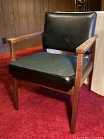 Photo of mid-century modern chair showing wooden frame, black vinyl upholstered backrest, and black fabric seat cushion against red carpet and wood paneled wall.