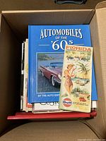 Top view of a box containing a hardcover book titled 'Automobiles of the '60s' along with other booklets and brochures including a Florida Gulf tourism vacation map