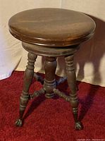 Photo of the vintage wood piano stool showing round wooden seat, turned legs, and metal claw feet on red carpet.