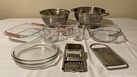 Photo displaying a large stainless-steel colander, a set of four stainless steel mixing bowls stacked together, a square Pyrex casserole dish with red handles, a round Anchor Hocking glass casserole dish, two round Anchor Hocking glass pie plates, Pyrex and Fire King glass measuring cups, a stainless steel box grater, and a stainless steel spaetzle noodle maker arranged neatly on a white surface.