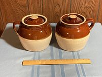 Side by side photo of two ceramic bean pots with lids, showing their size relative to ruler in front.