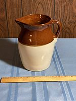 Photo of ceramic two-tone brown and cream pitcher with handle and spout on table, showing size against ruler