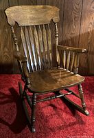 Photo shows a single wooden rocking chair with spindled back and contoured wooden seat in a medium brown finish against a wood paneled wall.