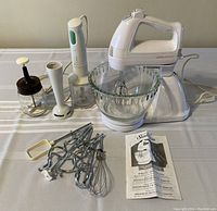 Photo showing white Sunbeam Mixmaster stand mixer with glass mixing bowl, multiple beaters, whisks and dough hooks laid out, a white immersion blender with chopping attachment, and a glass hand chopper with white lid.