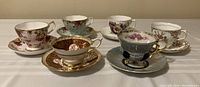 Five teacups with matching saucers displayed side by side on a white surface, showing a variety of floral and gold accented designs.