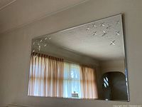 Photo of a large rectangular wall mirror with bevelled edges and star patterns etched into the upper corners, hanging on a wall reflecting a window with curtains.