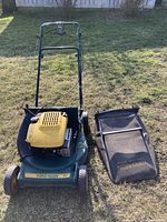 Photo showing yellow Yardman lawn mower next to a black leaf bag on grass outdoors. The mower has a yellow engine cover and green base with black handles and wheels.