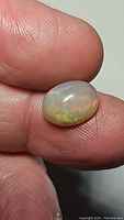 Close-up of the Ethiopian opal held between fingers showing the rainbow play of colors and smooth oval cabochon cut.