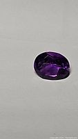 Photo of the oval cut deep purple Brazilian amethyst gemstone on a white background, showing facet reflections and color.