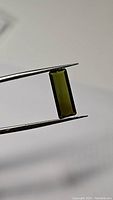 Close-up photo of the rectangular green tourmaline gemstone held by tweezers, showing color and clarity clearly