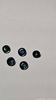 Five round cabochon deep blue sapphires with teal green tones, arranged on a white background showing translucency and smooth surface.
