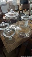 The full set on a wooden surface showing four clear glass jars of different sizes each with an attached lid and one clear glass bowl.