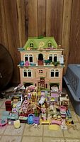 Front view of the pink Victorian doll house with green roof, multiple windows, and many accessories including figures, chairs, beds, and toys arranged in front on floor.