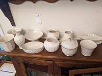 Wide view of all nine Lenox white porcelain pieces displayed on wooden furniture, showing planters, vases, vegetable dish, pedestal dish, covered candy dish and butter plate.