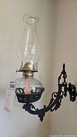 Close-up of one oil lamp wall sconce showing glass hurricane chimney, round clear glass oil reservoir with fabric wick, and black wrought iron ornate bracket mount.